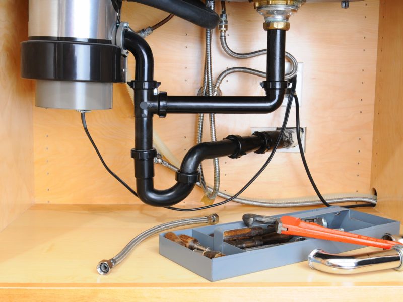 Detail of the plumbing system under a modern kitchen sink, with a plumbers tool tray and equipment. Horizontal format.