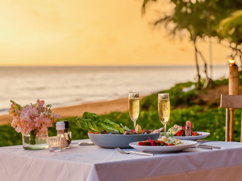Romantic sunset dinner on beach. Table honeymoon set with luxurious food, glasses of champagne drinks in restaurant with sea view and palm trees on background. Summer vacation or wedding concept.